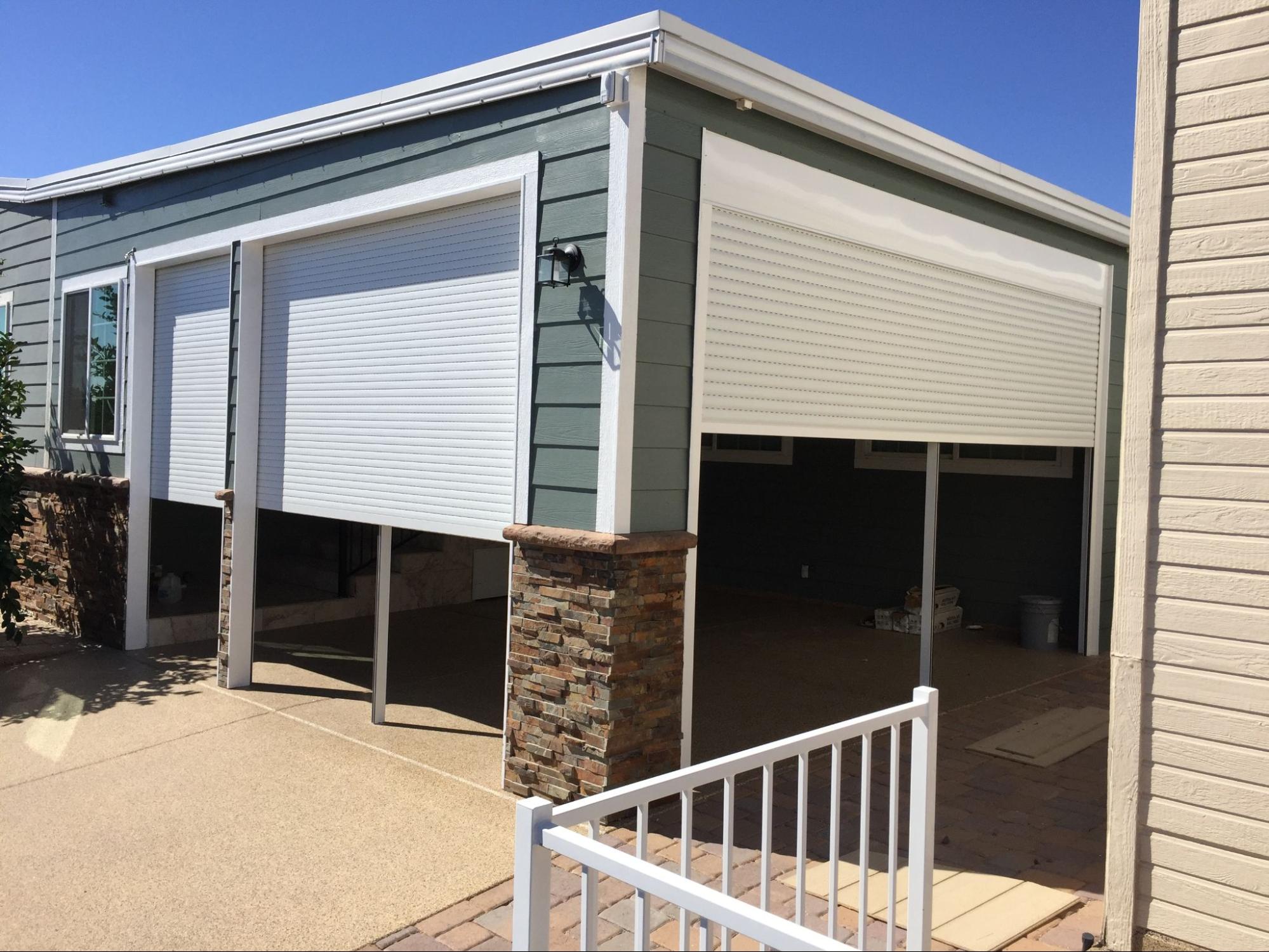 A modern garage with green siding and white rolling doors is partially open. Stone pillars add contrast. The scene is bright and sunny with blue skies.