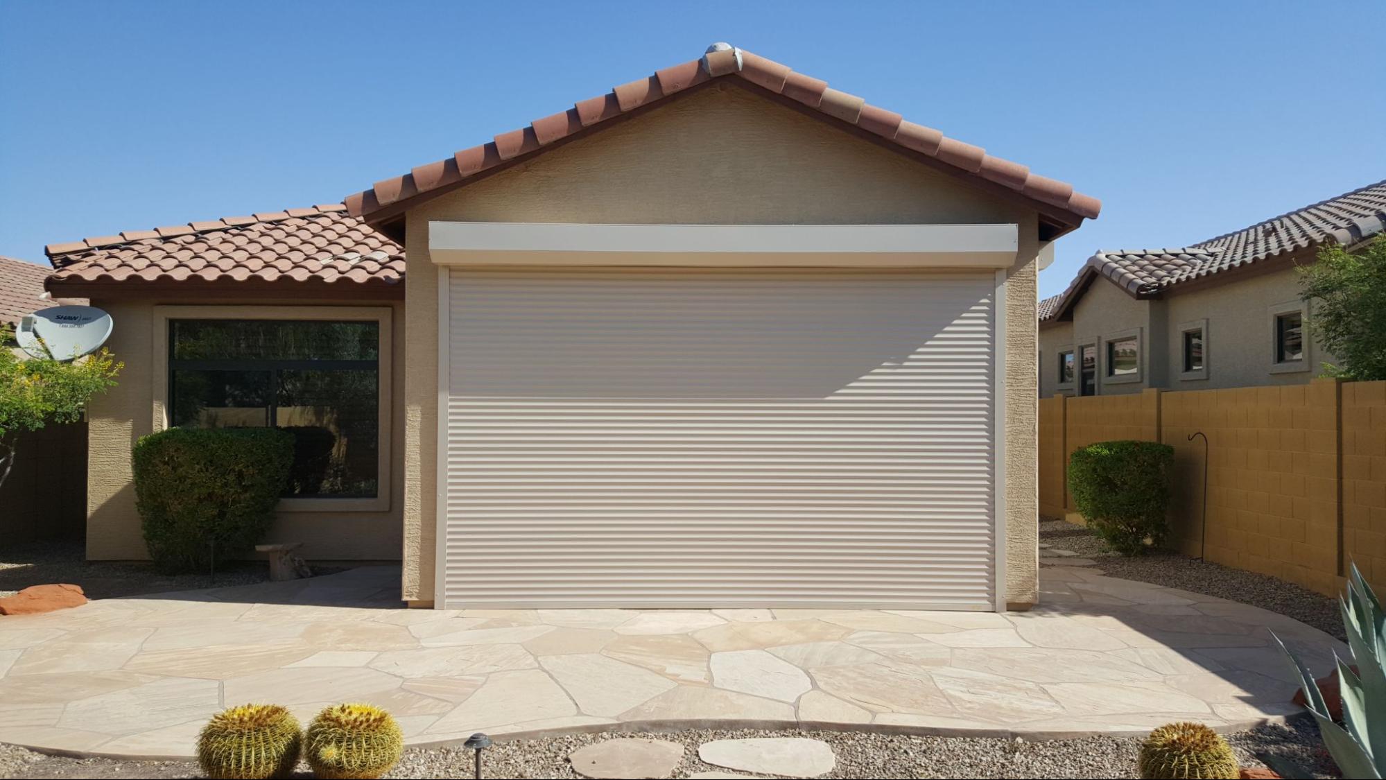 Single-story house with a closed beige garage door, terracotta roof tiles, surrounded by desert landscaping with cacti and stone pathway under clear blue sky.