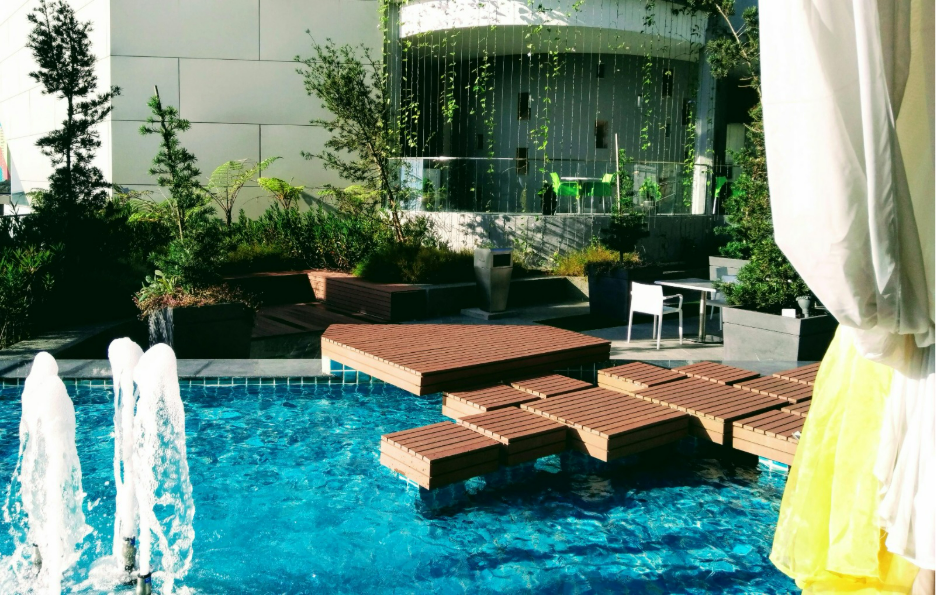 A serene outdoor scene features a blue pool with wooden platforms and water fountains. There's lush greenery, a modern building, and a relaxed ambiance.