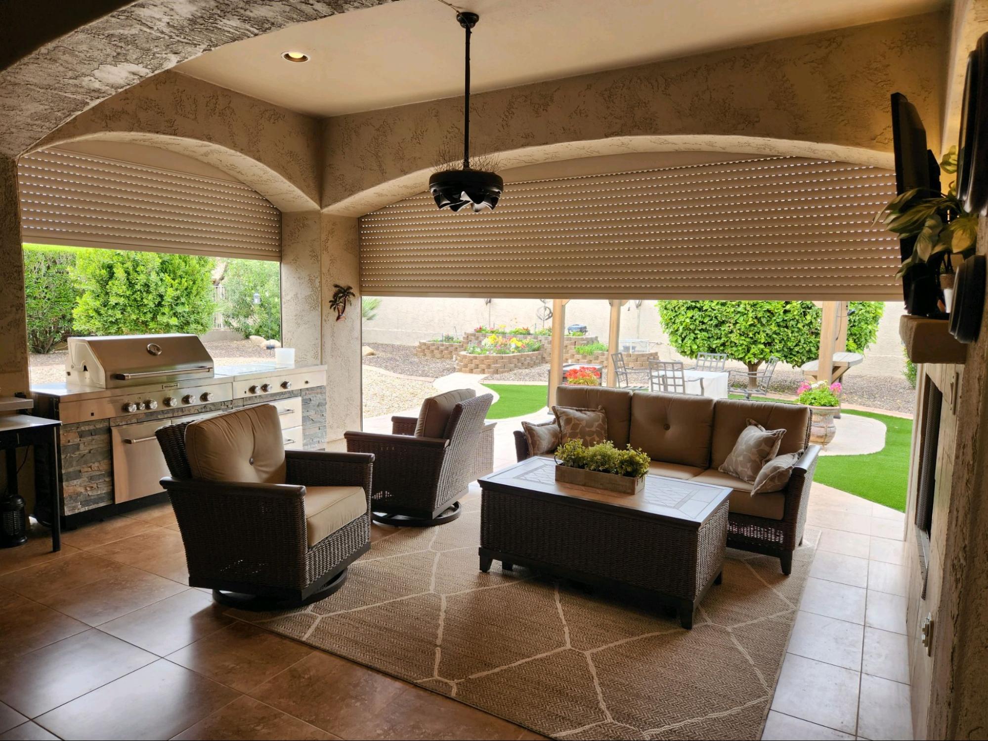 Cozy outdoor patio with wicker furniture, a grill, and a rug. Large window with rolled-up blinds opens to a lush garden and seating area, creating a warm ambiance.